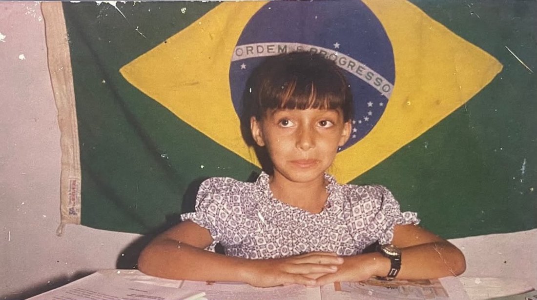 Patrícia Feitosa ainda menina, sentada à mesa com a bandeira do Brasil — origem no interior do Nordeste